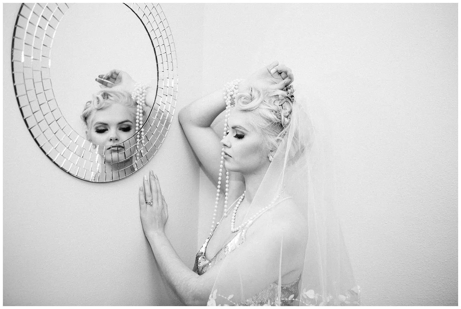Premier Snohomish & Seattle Wedding Photographer | GSquared Weddings Photography A bride in a veil and pearls stands by a wall, gently touching a round mosaic mirror, her reflection visible as she gazes downward with a thoughtful expression. This black and white moment captures timeless Snohomish Seattle wedding elegance. 47.9129° N, 122.0982° W | Serving Snohomish, Seattle , Orlando & Beyond