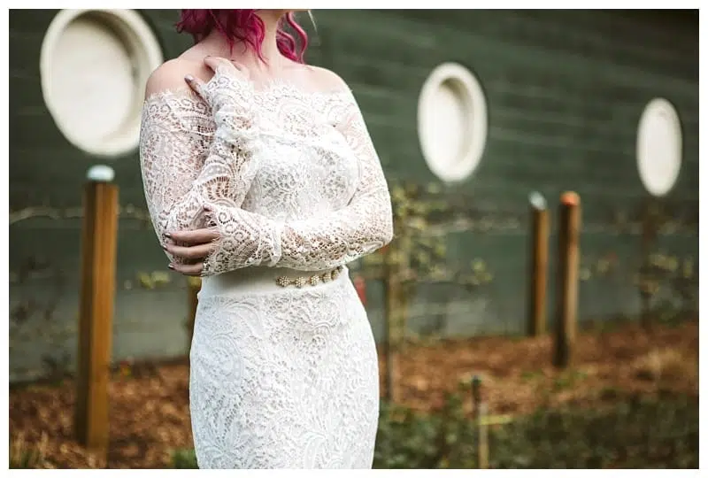 Premier Snohomish & Seattle Wedding Photographer | GSquared Weddings Photography A woman with pink hair wears an off-the-shoulder, long-sleeved lace wedding dress, standing outdoors near a green wall with round windows at her Snohomish Seattle wedding, with a garden in the background. 47.9129° N, 122.0982° W | Serving Snohomish, Seattle , Orlando & Beyond