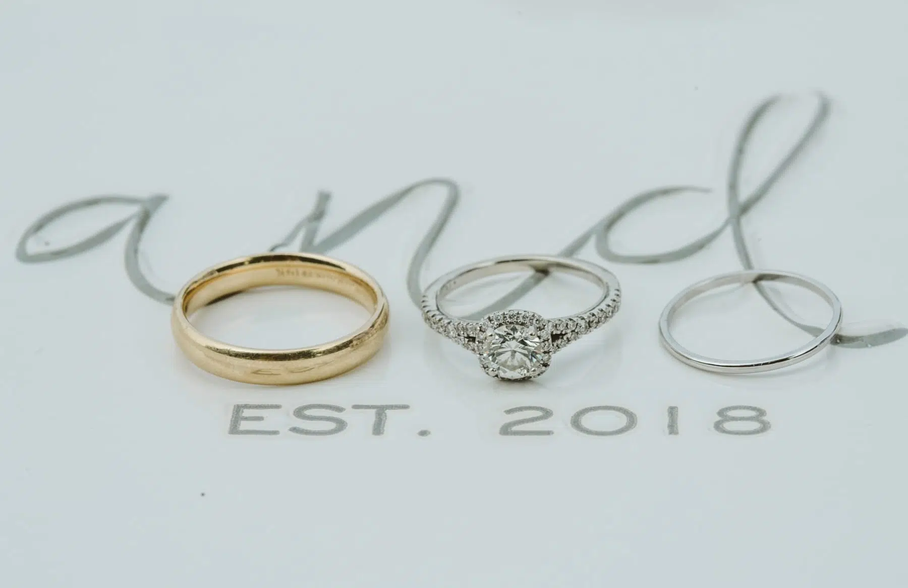 Three rings—a gold band, a diamond engagement ring, and a silver band—are arranged on a white surface over the words “EST. 2018” in gray calligraphy, capturing the timeless elegance of a Snohomish love story. Snohomish Wedding Photography