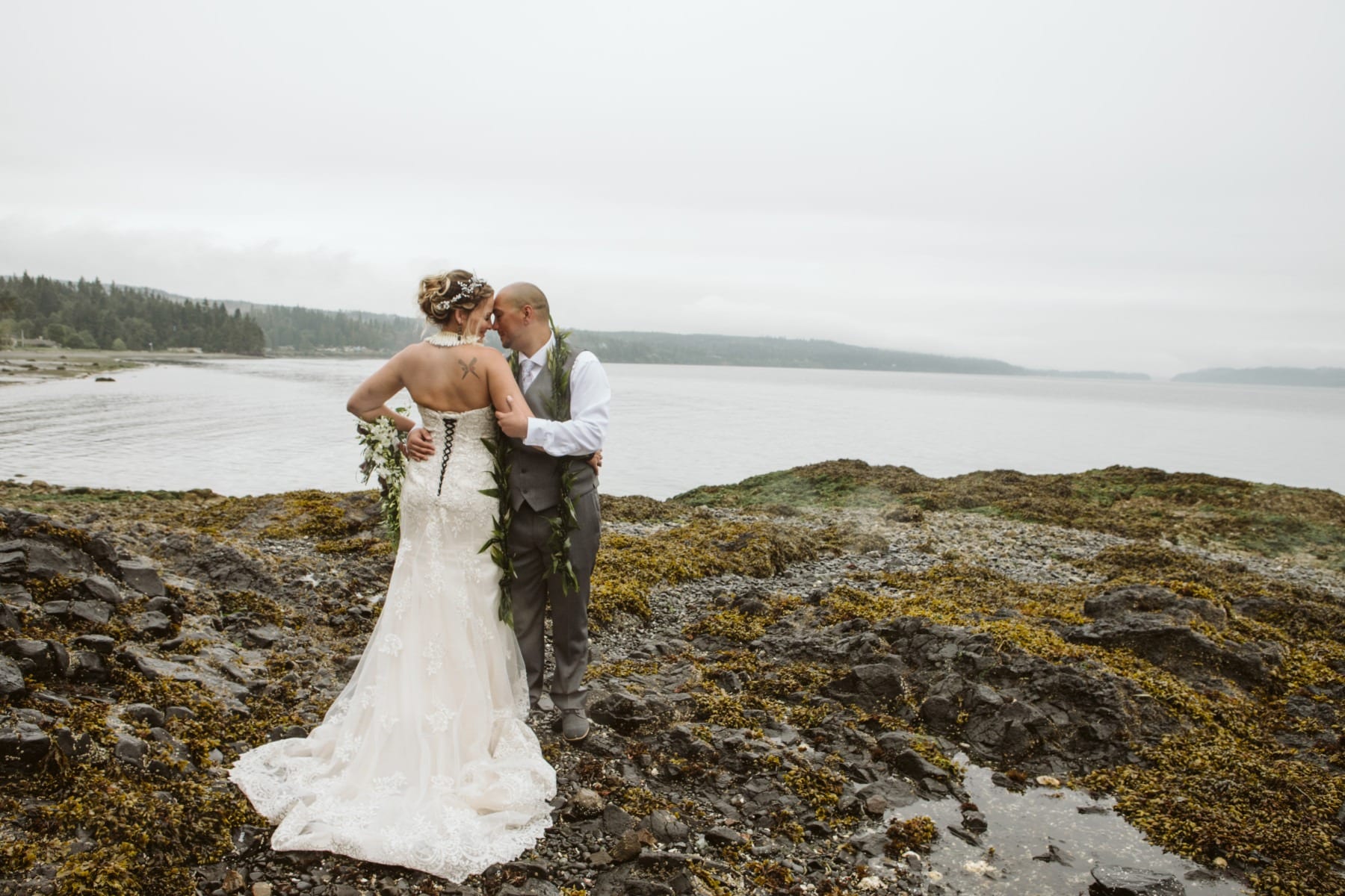 A bride and groom stand on a rocky, seaweed-covered Snohomish shore, embracing and kissing, with calm water and a misty forested coastline in the background. The bride wears a white gown and the groom is in a vest and tie. Snohomish Wedding Photography