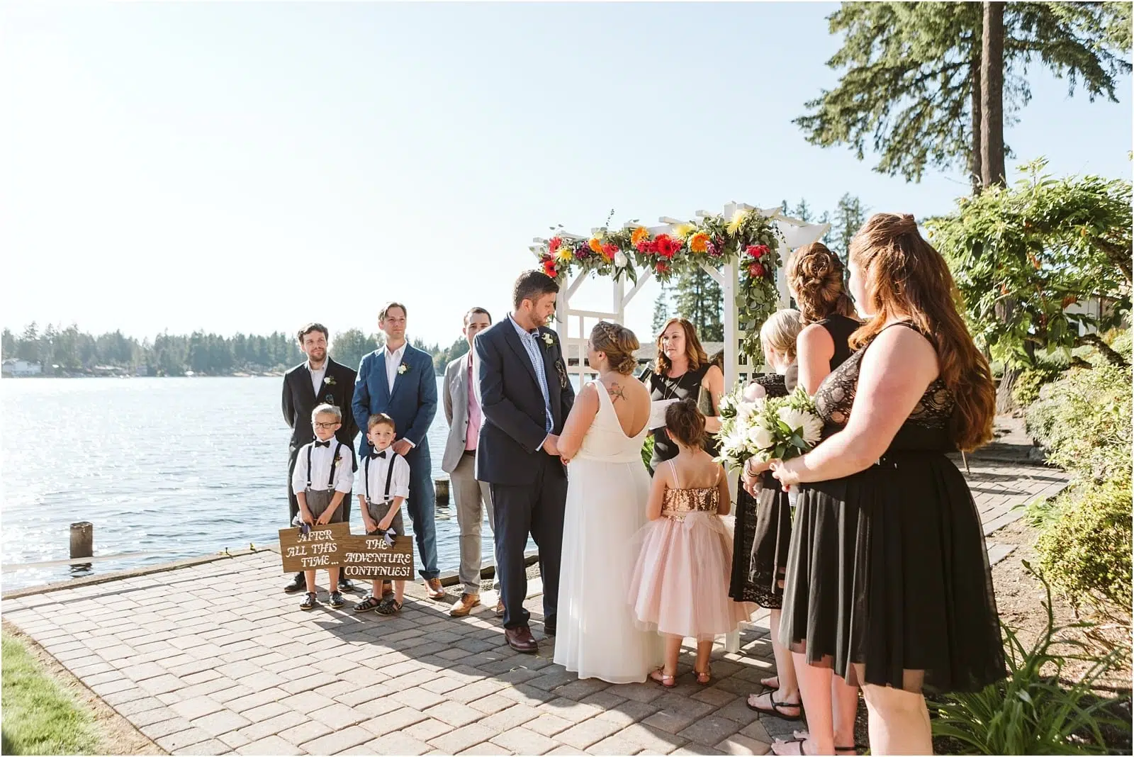 Premier Snohomish & Seattle Wedding Photographer | GSquared Weddings Photography A bride and groom stand under a flower-adorned arch by a lake, holding hands during their sunny Snohomish wedding ceremony. Family and friends watch as two children hold signs, celebrating this beautiful Seattle engagement. 47.9129° N, 122.0982° W | Serving Snohomish, Seattle , Orlando & Beyond