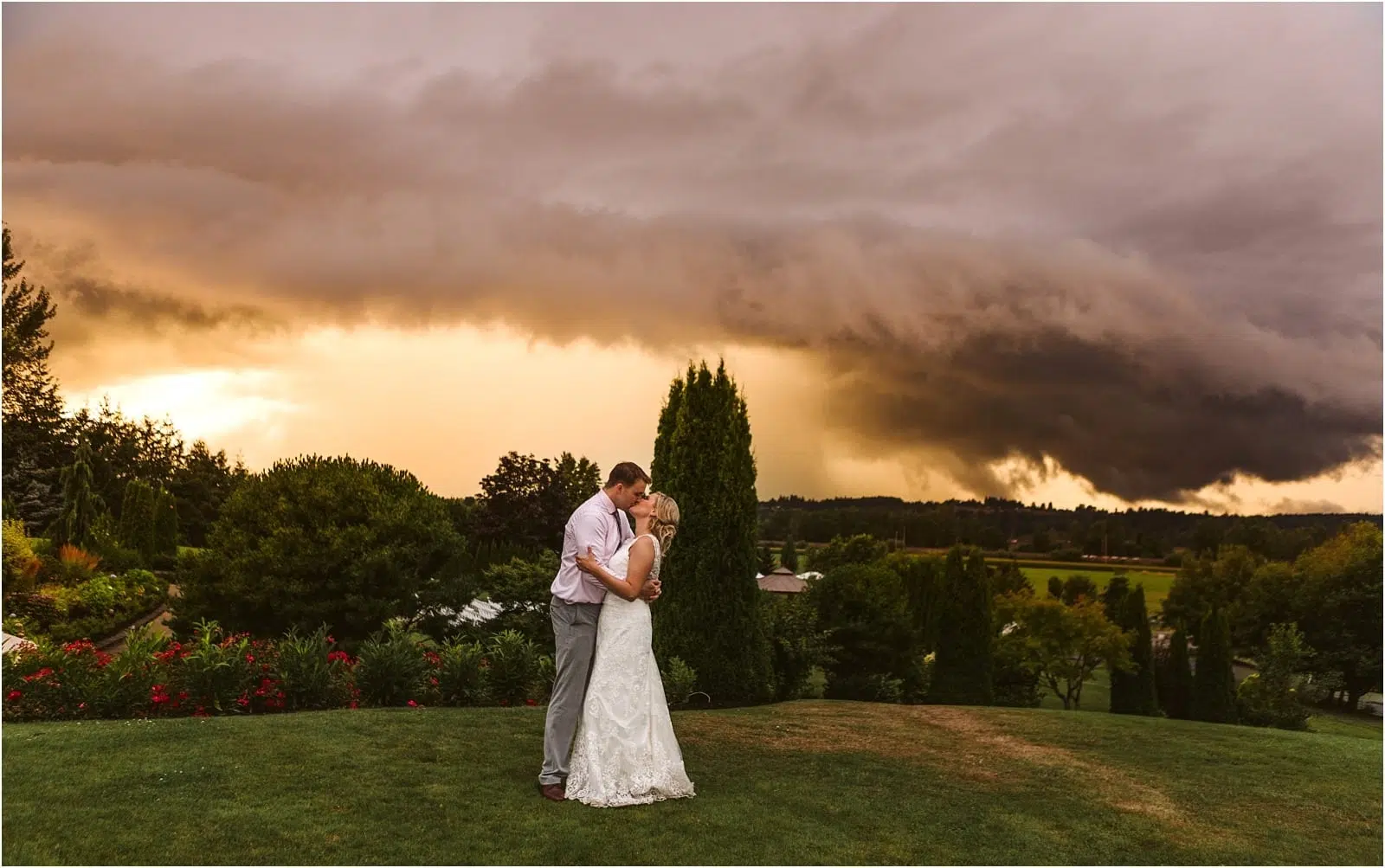 A bride and groom embrace on a grassy Snohomish hill at sunset, with dramatic dark storm clouds and golden light behind them, surrounded by lush trees and a scenic landscape. Snohomish Wedding Photography