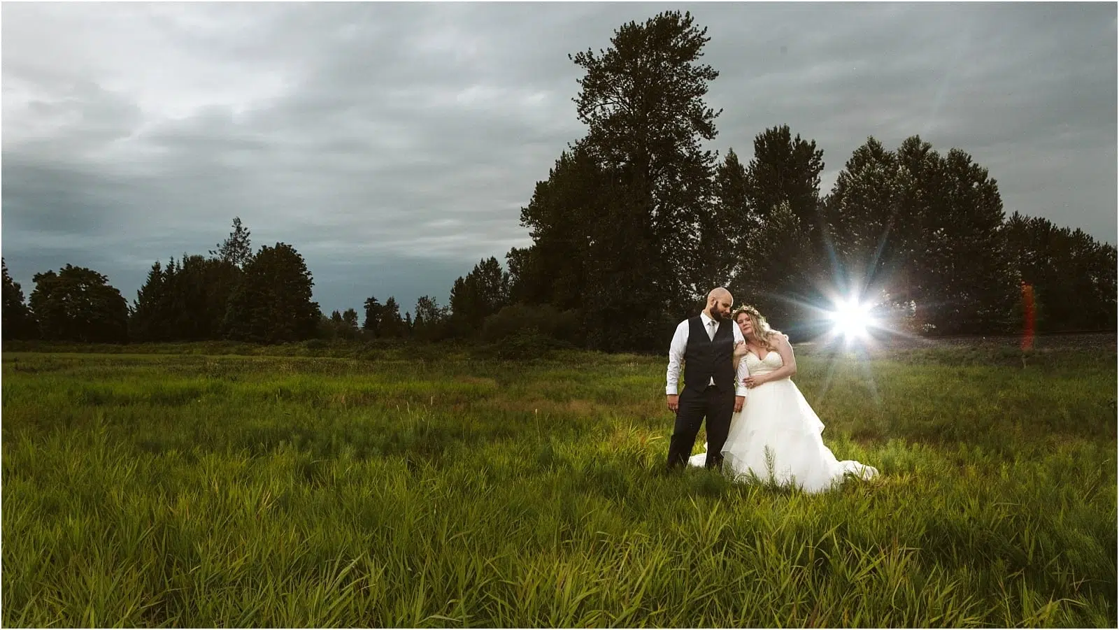 Premier Snohomish & Seattle Wedding Photographer | GSquared Weddings Photography A bride and groom stand together in a green field at dusk near Snohomish, with trees and a cloudy sky in the background. The bride leans on the groom, while a bright flash of light appears behind them during their Seattle wedding. 47.9129° N, 122.0982° W | Serving Snohomish, Seattle , Orlando & Beyond