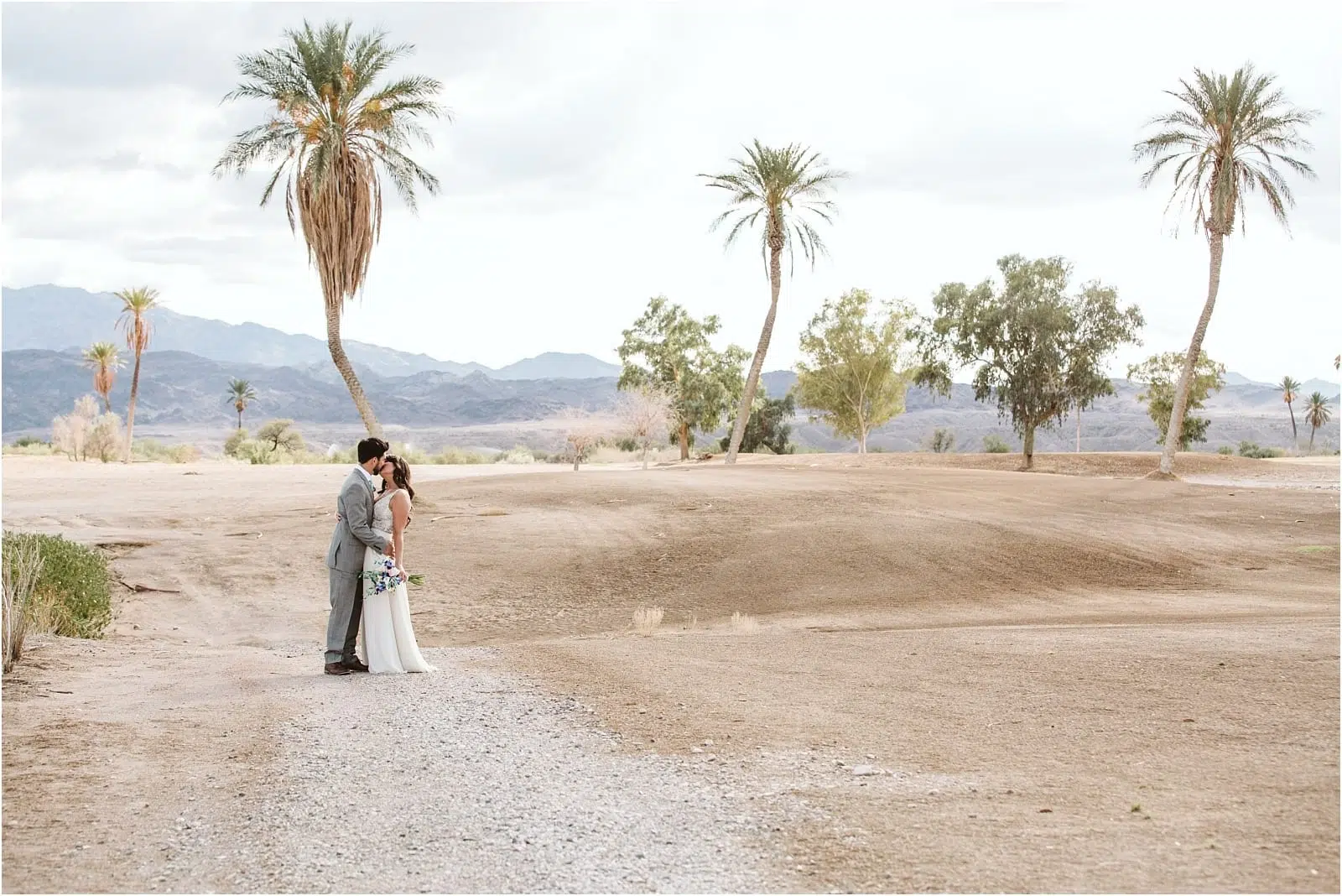 Premier Snohomish & Seattle Wedding Photographer | GSquared Weddings Photography A bride and groom stand close together, embracing on a dirt path surrounded by palm trees in a desert landscape with mountains in the background under a cloudy sky, evoking the romance of a Snohomish Seattle wedding engagement. 47.9129° N, 122.0982° W | Serving Snohomish, Seattle , Orlando & Beyond
