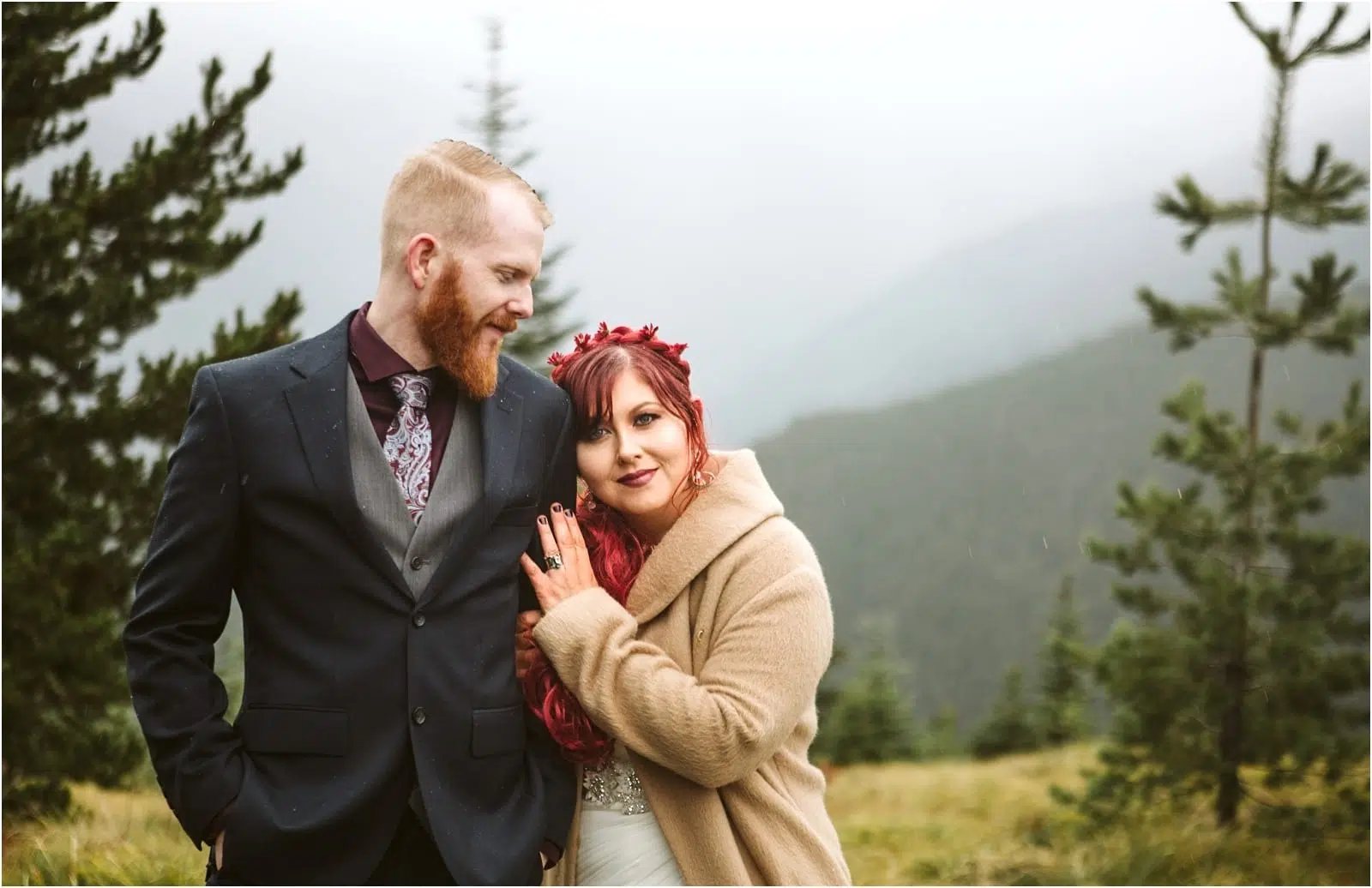 Premier Snohomish & Seattle Wedding Photographer | GSquared Weddings Photography A couple poses outdoors in front of misty mountains and pine trees for their Snohomish engagement. The man wears a dark suit and tie, while the woman, with red hair and a flower crown, smiles as she rests her head on his shoulder. 47.9129° N, 122.0982° W | Serving Snohomish, Seattle , Orlando & Beyond