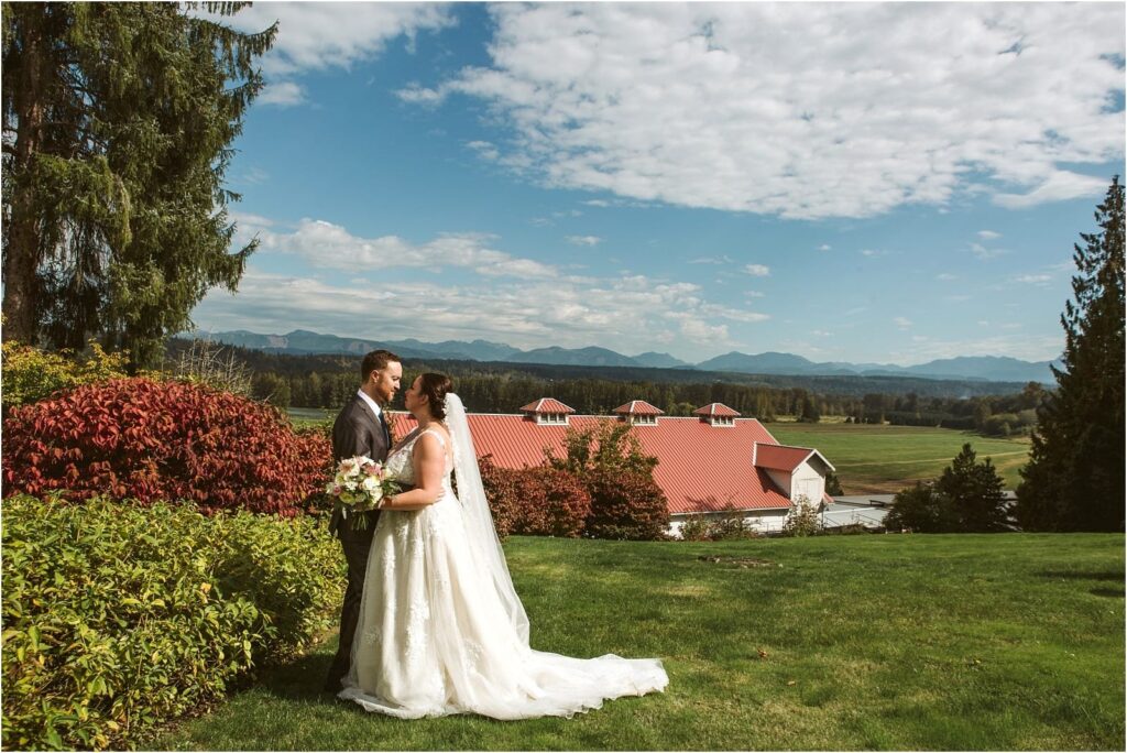 Premier Snohomish & Seattle Wedding Photographer | GSquared Weddings Photography A bride and groom stand facing each other on a grassy lawn in Snohomish, surrounded by greenery, with a red-roofed barn, open fields, and mountains in the background under a partly cloudy sky—a perfect Seattle wedding setting. 47.9129° N, 122.0982° W | Serving Snohomish, Seattle , Orlando & Beyond