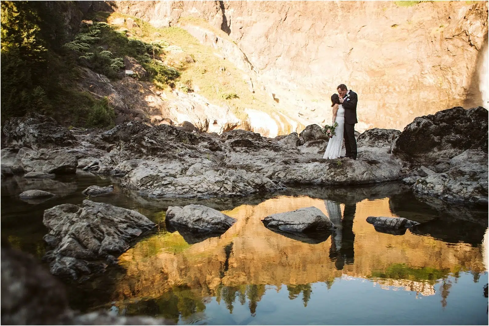 Premier Snohomish & Seattle Wedding Photographer | GSquared Weddings Photography A couple in wedding attire embrace on rocky terrain beside a mountain stream near Snohomish, with the golden cliffs and trees reflected in the calm water below—a breathtaking Seattle wedding engagement moment. 47.9129° N, 122.0982° W | Serving Snohomish, Seattle , Orlando & Beyond
