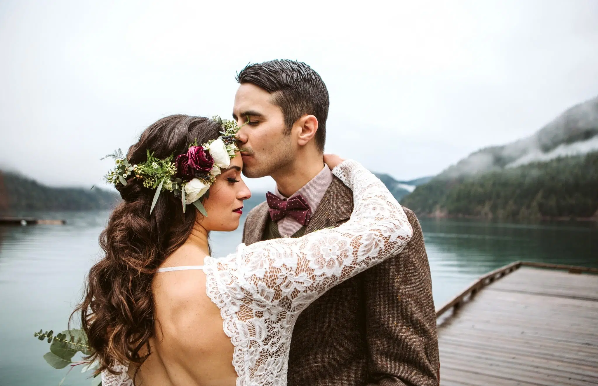 Premier Snohomish & Seattle Wedding Photographer | GSquared Weddings Photography A bride in a lace dress and floral crown embraces a groom in a suit and bow tie on a dock by the lake, misty forested mountains behind them. The groom kisses her forehead. Captured by GSquared Weddings, serving Snohomish & beyond. 47.9129° N, 122.0982° W | Serving Snohomish, Seattle , Orlando & Beyond