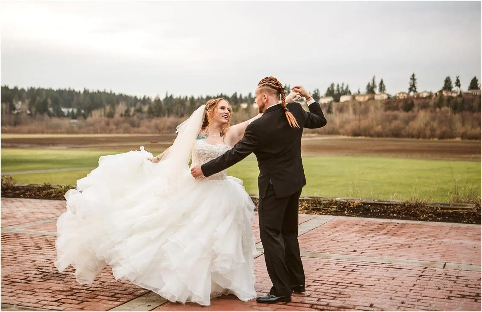 Premier Snohomish & Seattle Wedding Photographer | GSquared Weddings Photography A bride in a white gown and veil twirls joyfully with her groom in a black suit on a brick patio, capturing a magical Snohomish Seattle wedding moment amid grassy fields and trees under a cloudy sky. 47.9129° N, 122.0982° W | Serving Snohomish, Seattle , Orlando & Beyond