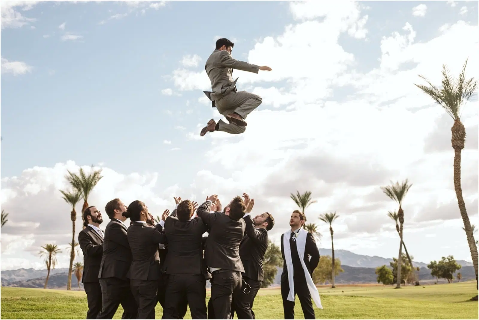 Premier Snohomish & Seattle Wedding Photographer | GSquared Weddings Photography A group of men in suits toss another man in a gray suit high into the air outdoors at a Snohomish Seattle wedding, surrounded by palm trees and mountains under a partly cloudy sky. 47.9129° N, 122.0982° W | Serving Snohomish, Seattle , Orlando & Beyond