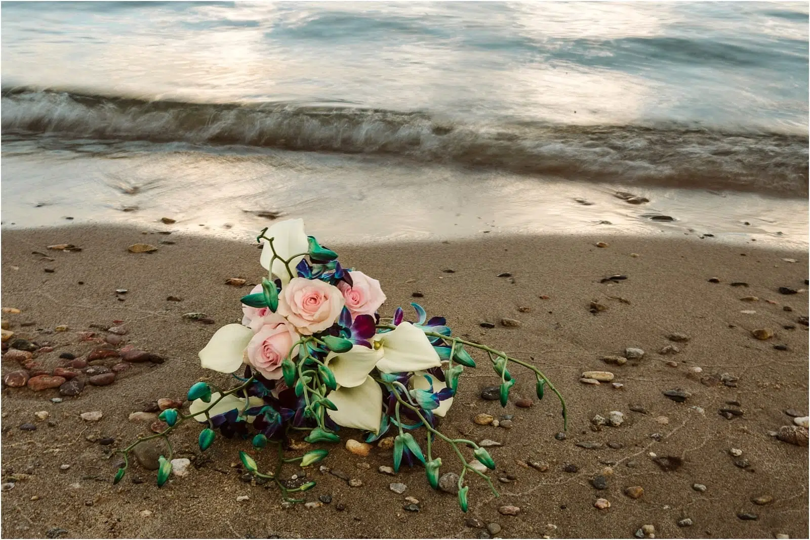 Premier Snohomish & Seattle Wedding Photographer | GSquared Weddings Photography A bouquet with pink roses, white calla lilies, and blue-purple flowers lies on a sandy beach near gentle waves—perfect for a Snohomish or Seattle wedding engagement, surrounded by small pebbles. 47.9129° N, 122.0982° W | Serving Snohomish, Seattle , Orlando & Beyond