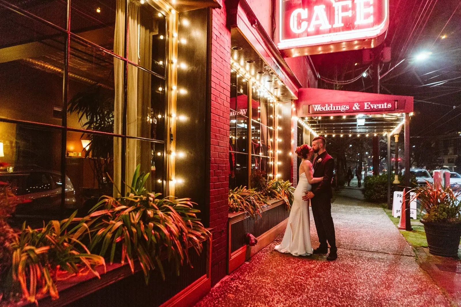 A couple in wedding attire kisses on a sidewalk outside a warmly lit café in Snohomish at night, beneath a glowing red sign that reads CAFE and Weddings & Events. Plants line the windows and street. Snohomish Wedding Photography