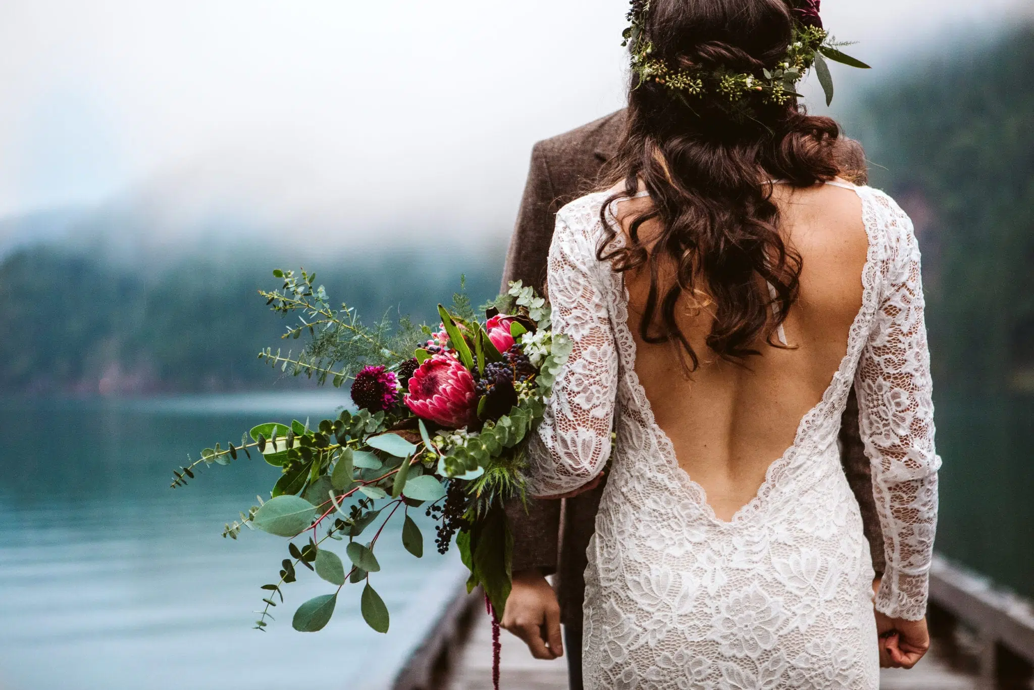 A bride with long wavy hair and a floral crown stands on a dock in Snohomish, holding a bouquet of flowers. She wears a lace dress with an open back. A groom stands partly hidden behind her, with a lake and misty forest in the background. Snohomish Wedding Photography