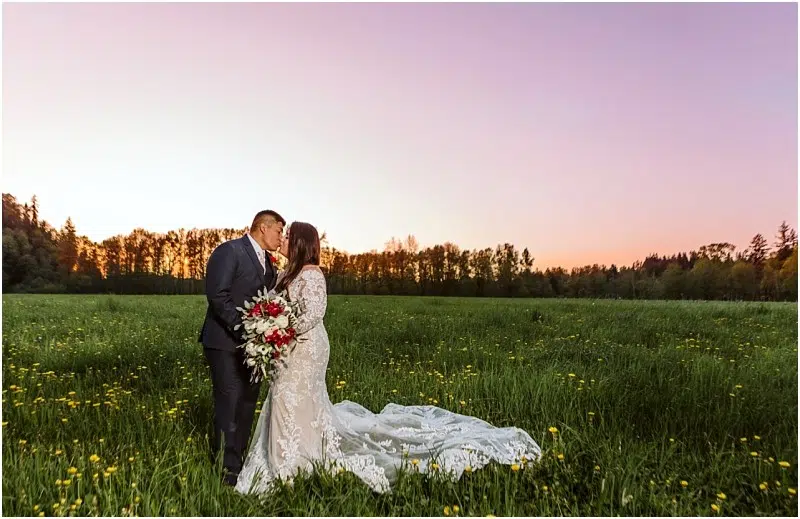Premier Snohomish & Seattle Wedding Photographer | GSquared Weddings Photography A bride and groom stand in a green field at sunset, kissing during their Snohomish wedding. The bride wears a long white lace dress with a train and holds a bouquet of red and white flowers. Trees line the horizon. 47.9129° N, 122.0982° W | Serving Snohomish, Seattle , Orlando & Beyond