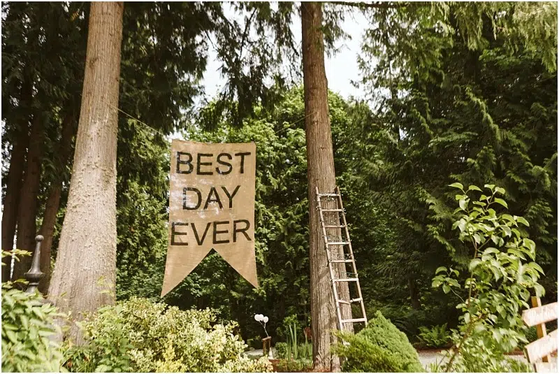Premier Snohomish & Seattle Wedding Photographer | GSquared Weddings Photography A large burlap banner reading BEST DAY EVER hangs between two tall trees in a lush, green forested area—an idyllic scene perfect for a Snohomish Seattle wedding or engagement. A wooden ladder leans against one of the trees nearby. 47.9129° N, 122.0982° W | Serving Snohomish, Seattle , Orlando & Beyond