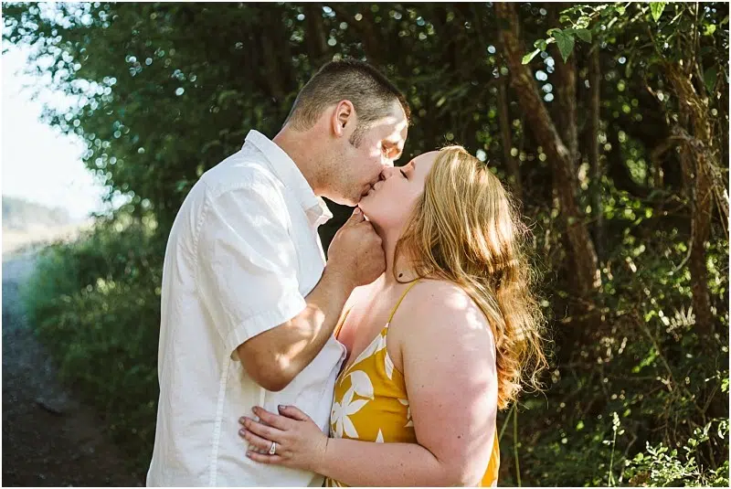 Premier Snohomish & Seattle Wedding Photographer | GSquared Weddings Photography A couple shares a kiss in a sunlit, wooded area during their Snohomish engagement session. The man wears a white shirt, and the woman, in a yellow dress with white floral patterns, glows in the romantic Seattle-area light. 47.9129° N, 122.0982° W | Serving Snohomish, Seattle , Orlando & Beyond