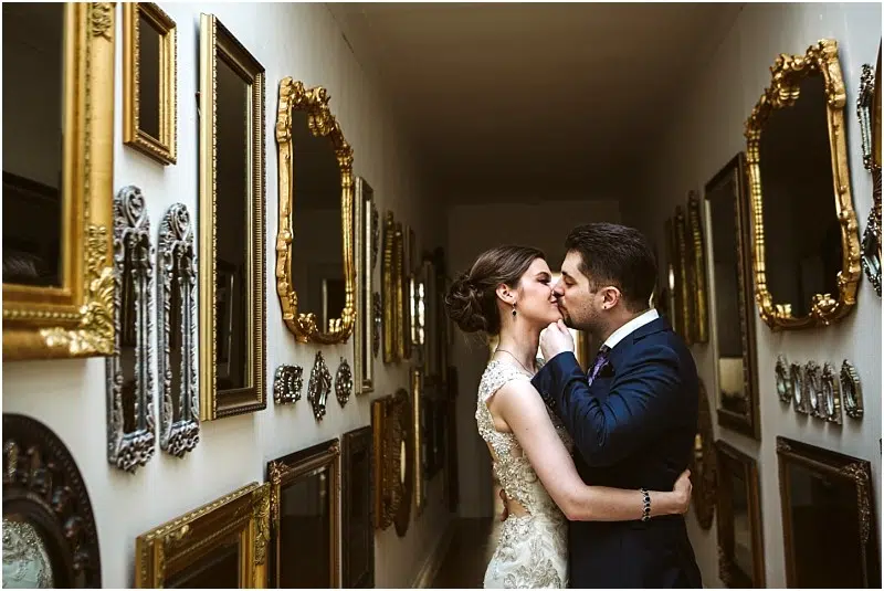 A bride and groom embrace and kiss in a narrow Snohomish hallway lined with ornate gold-framed mirrors and vintage picture frames, both dressed in formal wedding attire. Snohomish Wedding Photography