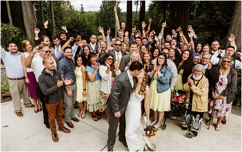 Premier Snohomish & Seattle Wedding Photographer | GSquared Weddings Photography A large group of wedding guests stands outdoors, smiling and cheering with raised hands, as a bride and groom kiss in front. The joyful scene unfolds amid trees and greenery at a beautiful Snohomish Seattle wedding. 47.9129° N, 122.0982° W | Serving Snohomish, Seattle , Orlando & Beyond
