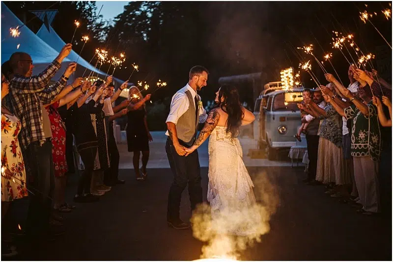 A newlywed couple holds hands and kisses while walking through a crowd of guests holding sparklers at dusk, celebrating outdoors in Snohomish with a decorated vintage van in the background. Snohomish Wedding Photography
