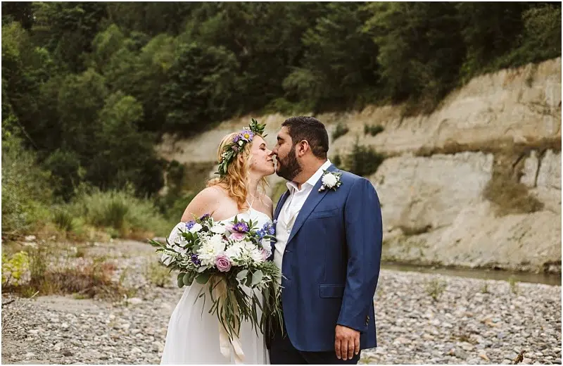 A bride in a white dress and flower crown kisses a groom in a blue suit, holding a bouquet, as they stand on a rocky Snohomish riverbank with a forested cliff in the background. Snohomish Wedding Photography