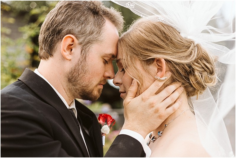 Premier Snohomish & Seattle Wedding Photographer | GSquared Weddings Photography A groom gently holds his bride’s face as they touch foreheads, eyes closed, in a tender moment. Captured outdoors during their Snohomish Seattle wedding, she wears a veil and necklace; he dons a black suit with a red boutonniere. 47.9129° N, 122.0982° W | Serving Snohomish, Seattle , Orlando & Beyond