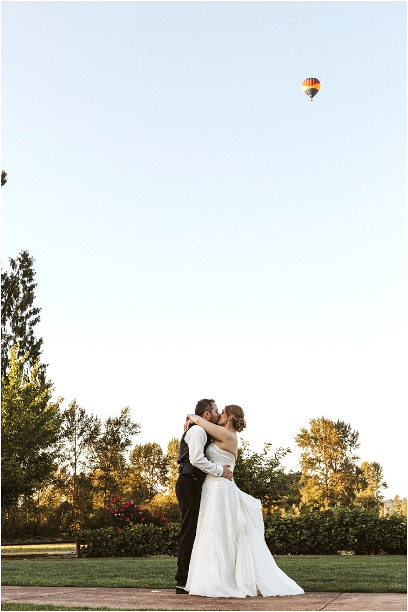 Premier Snohomish & Seattle Wedding Photographer | GSquared Weddings Photography A bride and groom embrace and kiss outdoors on a sunny day in Snohomish, surrounded by greenery, with a colorful hot air balloon floating in the clear blue sky above them—a dreamy Seattle wedding engagement moment. 47.9129° N, 122.0982° W | Serving Snohomish, Seattle , Orlando & Beyond