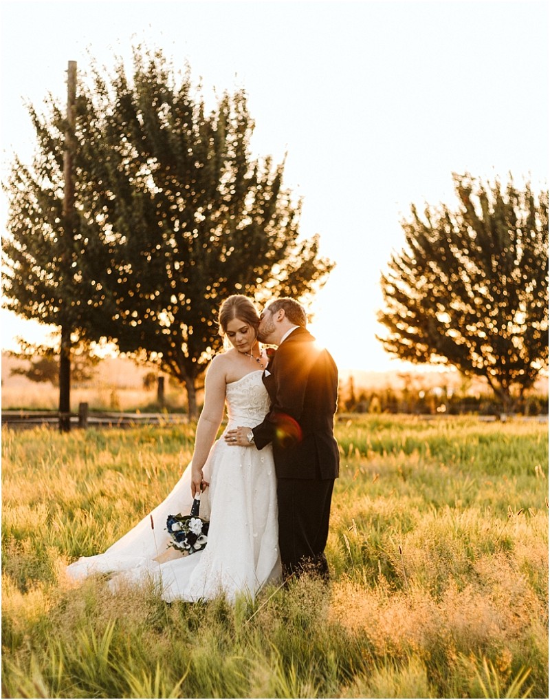 Premier Snohomish & Seattle Wedding Photographer | GSquared Weddings Photography A bride in a white gown holds a bouquet while the groom in a black suit kisses her cheek in a sunlit Snohomish field, with trees and golden light making their Seattle wedding engagement unforgettable. 47.9129° N, 122.0982° W | Serving Snohomish, Seattle , Orlando & Beyond