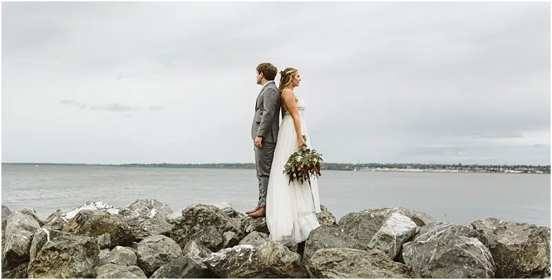 Premier Snohomish & Seattle Wedding Photographer | GSquared Weddings Photography A bride and groom stand back to back on large rocks by the water, facing opposite directions. The overcast sky and distant shoreline set a serene mood—perfect for a Snohomish Seattle wedding or engagement. The bride holds a bouquet of flowers. 47.9129° N, 122.0982° W | Serving Snohomish, Seattle , Orlando & Beyond
