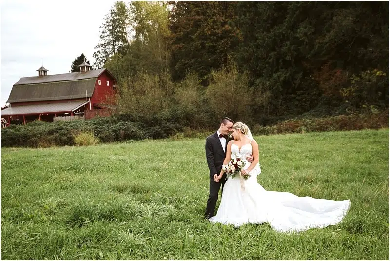 Premier Snohomish & Seattle Wedding Photographer | GSquared Weddings Photography A bride and groom stand together in a grassy field, holding hands and smiling, with a red barn and trees in the background. Perfect for a Snohomish or Seattle wedding engagement, the bride holds a bouquet and the groom wears a black suit. 47.9129° N, 122.0982° W | Serving Snohomish, Seattle , Orlando & Beyond