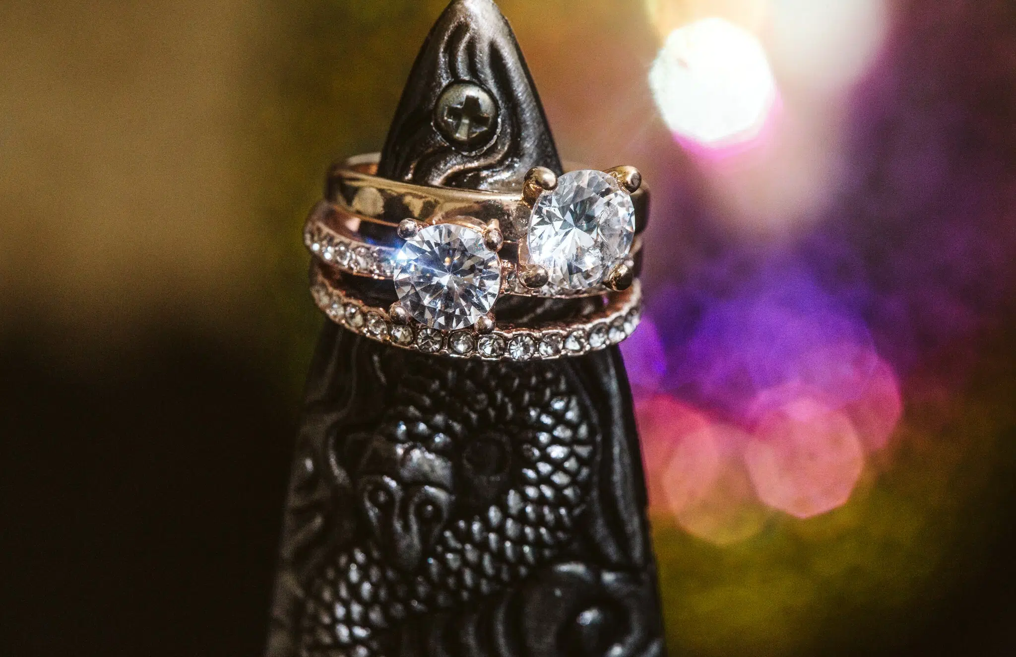 Snohomish & Seattle Wedding Photographer | GSquared Weddings Photography Two diamond rings—one with a row of small diamonds, another with two large stones—rest on a dark, intricately carved object featuring a dragon motif. Blurred colorful lights glow in the background. See more in The Weducation Series: 15 Things You Shouldn't Do on Your Wedding Day. 47.9129° N, 122.0982° W | wedding photo by GSquared Weddings