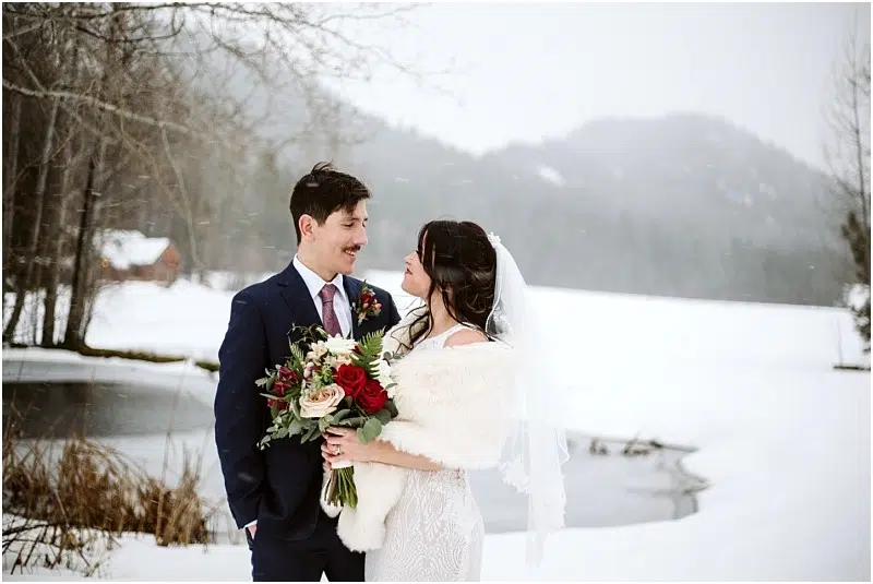 Snohomish & Seattle Wedding Photographer | GSquared Weddings Photography A bride and groom stand outdoors in the snow at their Leavenworth wedding, smiling at each other. The bride holds a red and white bouquet and wears a veil and fur shawl, while snowy trees and a pond complete the wintry scene. 47.9129° N, 122.0982° W | wedding photo by GSquared Weddings