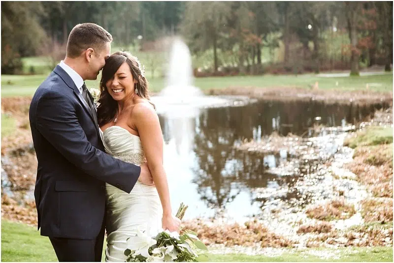 Snohomish & Seattle Wedding Photographer | GSquared Weddings Photography A bride and groom smile and embrace outdoors near a pond with a fountain on a lush golf course wedding, surrounded by greenery and trees. The bride holds a bouquet in her white dress while the groom wears a dark suit. 47.9129° N, 122.0982° W | wedding photo by GSquared Weddings