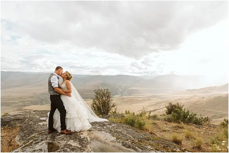 Snohomish & Seattle Wedding Photographer | GSquared Weddings Photography A bride and groom embrace and kiss on a rocky ledge during their Methow Elopement, overlooking a vast landscape of rolling hills beneath a cloudy sky. She wears a white gown, and he’s in a vest and dress shirt. 47.9129° N, 122.0982° W | wedding photo by GSquared Weddings