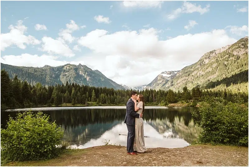 Snohomish & Seattle Wedding Photographer | GSquared Weddings Photography A couple stands embracing on the shore of Gold Creek Pond, surrounded by pine trees and mountains under a partly cloudy sky, their reflection mirrored in the calm lake—capturing a perfect engagement moment. 47.9129° N, 122.0982° W | wedding photo by GSquared Weddings
