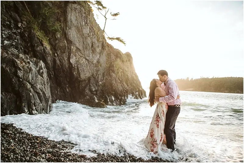 Snohomish & Seattle Wedding Photographer | GSquared Weddings Photography A couple embraces and kisses at the edge of the ocean during their Deception Pass Engagement Session in June 2020, standing in shallow waves by a rocky cliff at sunset. The woman wears a white floral dress; the man wears a light shirt and dark pants. 47.9129° N, 122.0982° W | wedding photo by GSquared Weddings