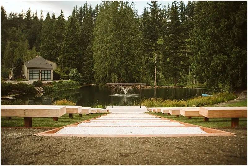 Snohomish & Seattle Wedding Photographer | GSquared Weddings Photography A wide stone path with wooden benches on both sides leads down to a small pond with a fountain and a graceful gray bridge, surrounded by tall trees and a house to the left. The scene is peaceful and green. 47.9129° N, 122.0982° W | wedding photo by GSquared Weddings