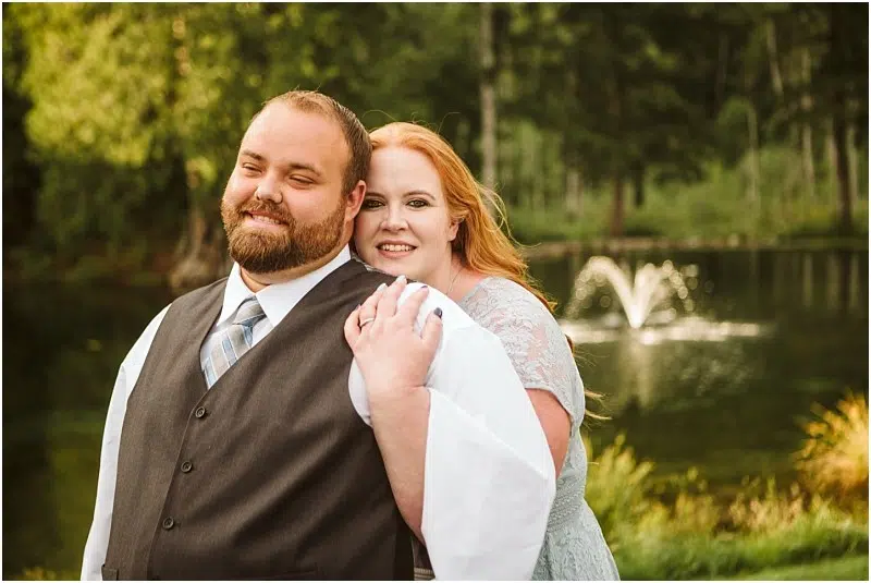 Snohomish & Seattle Wedding Photographer | GSquared Weddings Photography A smiling couple poses outdoors by a pond with a fountain for their Gray Bridge Engagement. The man wears a vest and tie while the woman, in a light blue dress, embraces him as trees and greenery fill the background. 47.9129° N, 122.0982° W | wedding photo by GSquared Weddings