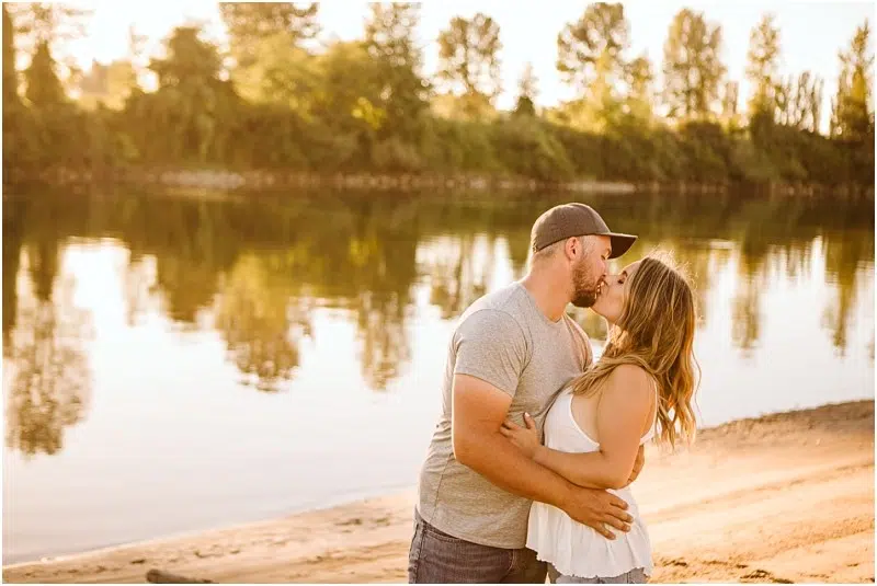 Snohomish & Seattle Wedding Photographer | GSquared Weddings Photography A couple kisses on a sandy riverbank at sunset during their Pemberton Farm engagement near Snohomish, with trees and their reflections visible in the calm water. The man wears a cap and grey shirt; the woman wears a white sleeveless top. 47.9129° N, 122.0982° W | wedding photo by GSquared Weddings