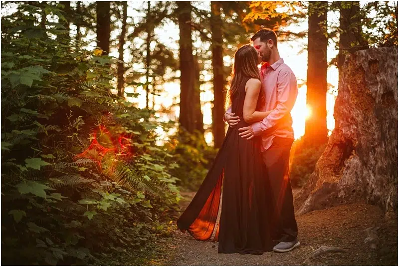 Snohomish & Seattle Wedding Photographer | GSquared Weddings Photography A couple embraces on a forest path at sunset during their Deception Pass August engagement. Sunlight streams through the trees, illuminating the woman's long black dress and casting a warm glow around them. 47.9129° N, 122.0982° W | wedding photo by GSquared Weddings