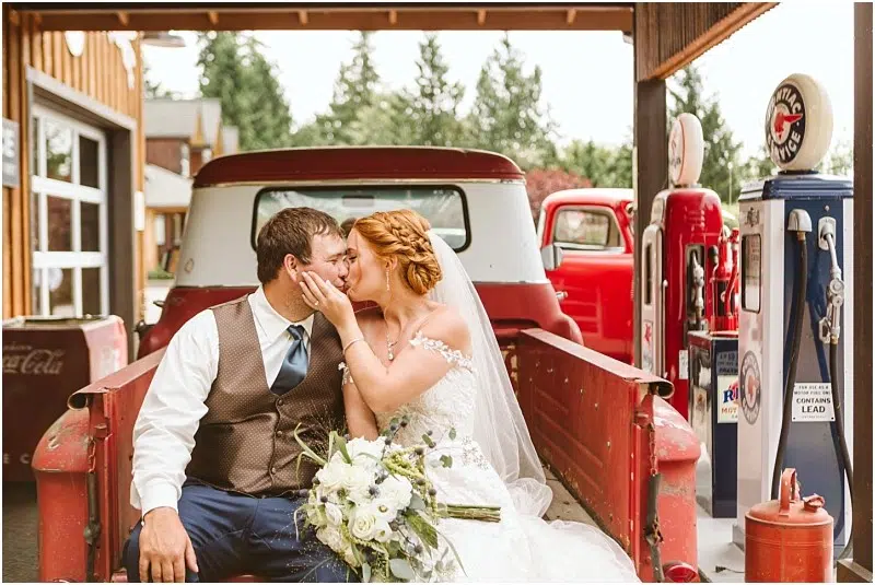 A bride and groom sit on the tailgate of a red vintage truck in Snohomish, sharing a kiss. The bride holds a white bouquet, with old-fashioned gas pumps and rustic buildings in the background. Snohomish Wedding Photography