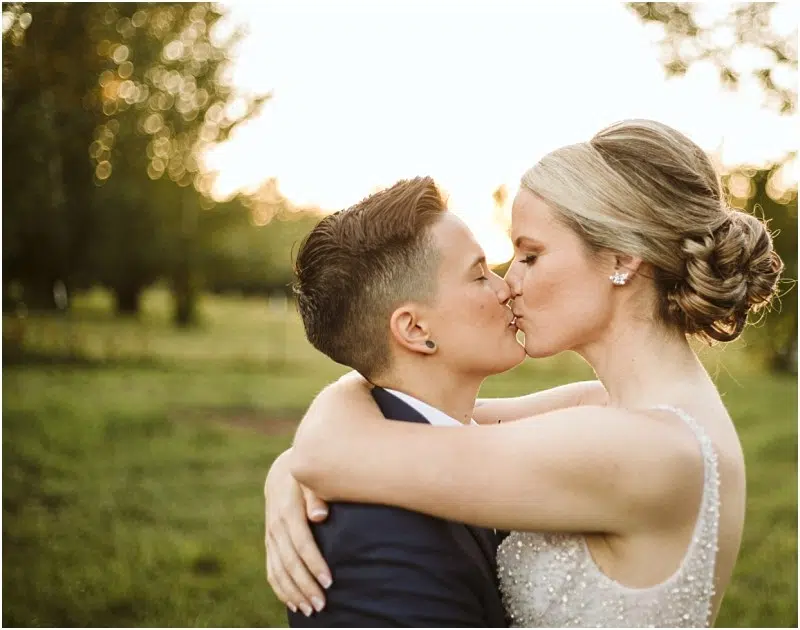 Two women embrace and kiss outdoors at sunset in Snohomish, one with short hair in a suit and the other with long hair in an updo and a beaded dress, surrounded by trees and soft sunlight. Snohomish Wedding Photography