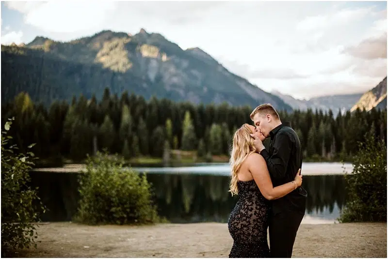 A couple embraces and kisses by a peaceful lake in Snohomish, surrounded by dense evergreen trees, with tall mountains and a partly cloudy sky in the background. Snohomish Wedding Photography