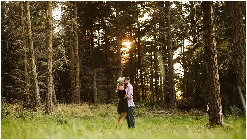 A couple embraces and kisses in a sunlit Snohomish forest clearing, surrounded by tall trees with golden sunlight streaming through the branches. The scene feels warm and romantic. Snohomish Wedding Photography