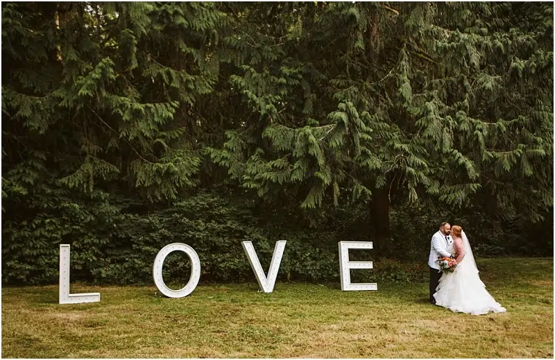 A bride and groom embrace and kiss outdoors in Snohomish, in front of large white letters spelling LOVE, with tall green trees in the background. Snohomish Wedding Photography