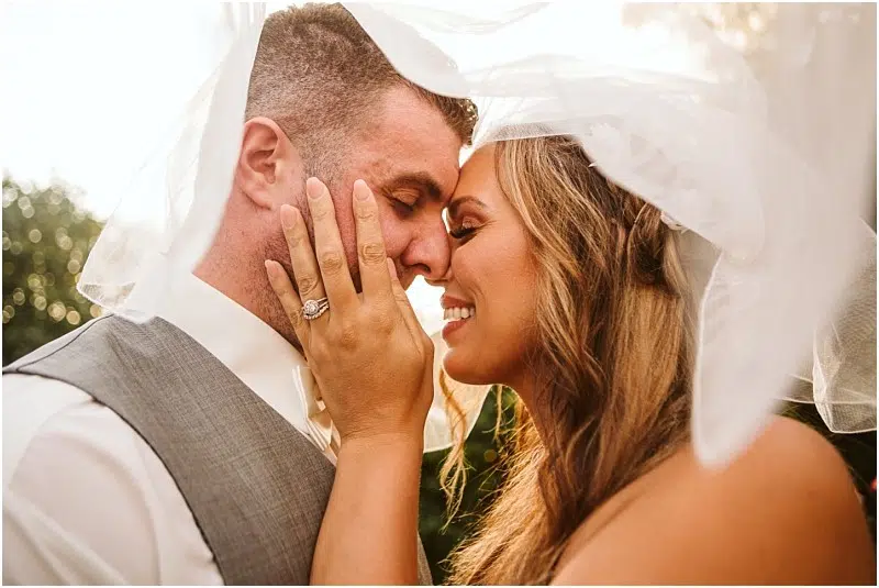 A bride and groom smile joyfully under a flowing veil in Snohomish, touching foreheads and holding each other close outdoors in warm, golden sunlight. The bride's hand, showing her ring, gently touches the groom's face. Snohomish Wedding Photography