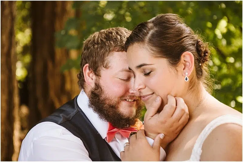 A couple embraces outdoors in Snohomish; the man with a beard and red bow tie gently holds the woman’s face as they smile with closed eyes, surrounded by trees and greenery in soft sunlight. Snohomish Wedding Photography