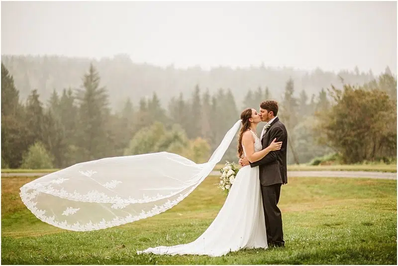 A bride and groom kiss outdoors on a grassy Snohomish field, surrounded by trees. The bride’s long lace veil flows dramatically in the wind as she holds a bouquet and the groom embraces her. Snohomish Wedding Photography