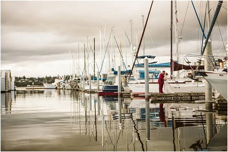 A Snohomish marina scene with several docked sailboats on calm water. Two people in red stand on a dock beside a white sailboat, with reflections of boats and masts shimmering under a cloudy sky. Snohomish Wedding Photography