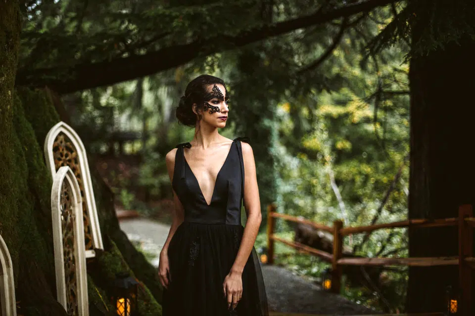 A woman in a black dress with a deep neckline and black lace mask stands outdoors on a forested path in Snohomish, surrounded by lush greenery and wooden railings, looking off to the side. Snohomish Wedding Photography