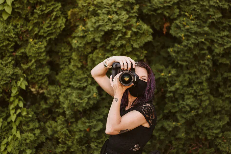 A person with purple hair and a black face mask holds a camera up to their eye, taking a photo. Standing in front of a lush green hedge in Snohomish, they wear a black sleeveless top. Snohomish Wedding Photography